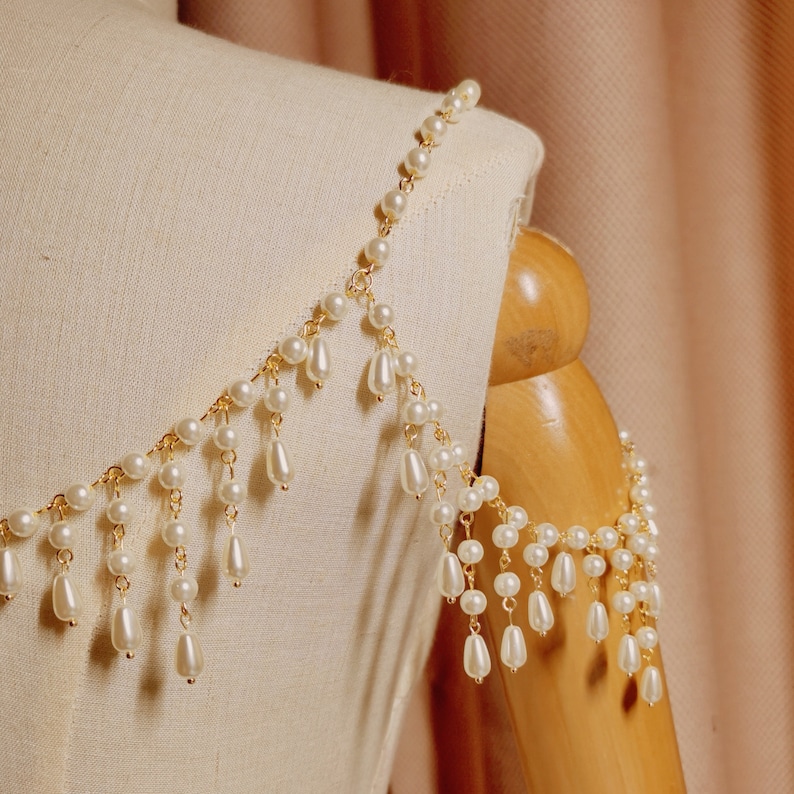 Op de afbeelding: Een gouden ketting met witte parels en hangende pareldruppels. De ketting is over een wit stoffen mannequin gelegd.