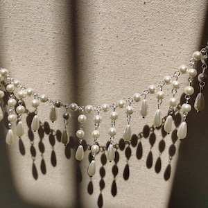 Op de afbeelding: Een witte parelketting met een franje van parels die eronder hangen. De ketting ligt op een witte stoffen achtergrond.