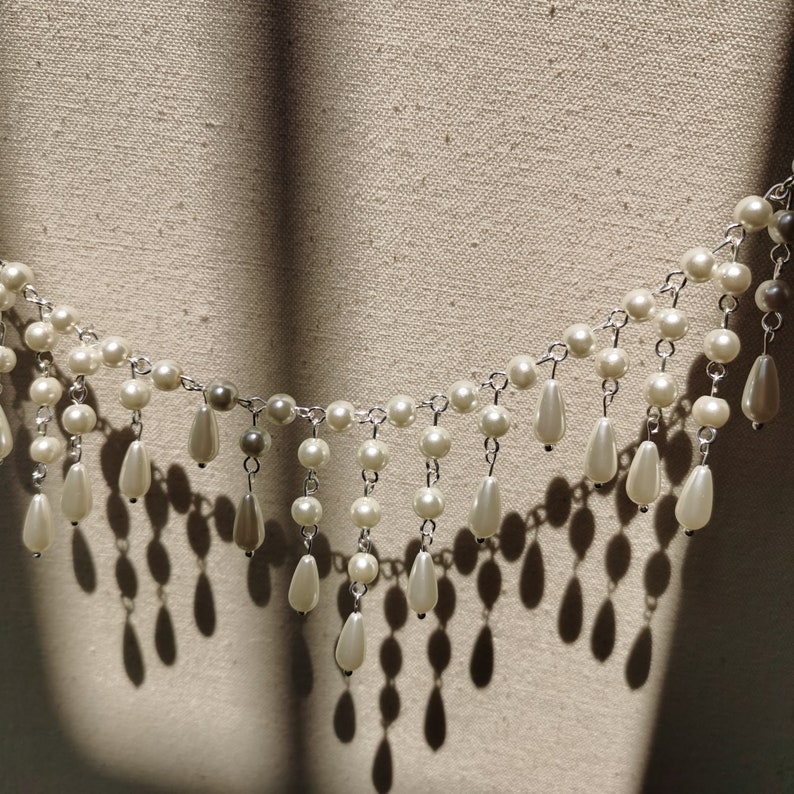 Op de afbeelding: Een witte parelketting met een franje van parels die eronder hangen. De ketting ligt op een witte stoffen achtergrond.