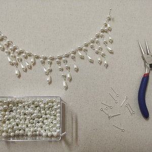 Op de afbeelding: Een close-up van een parelketting met een traanvormig ontwerp, een container met losse parels, een tang en een stapel springringen. De ketting is gemaakt van witte parels en een zilveren ketting.