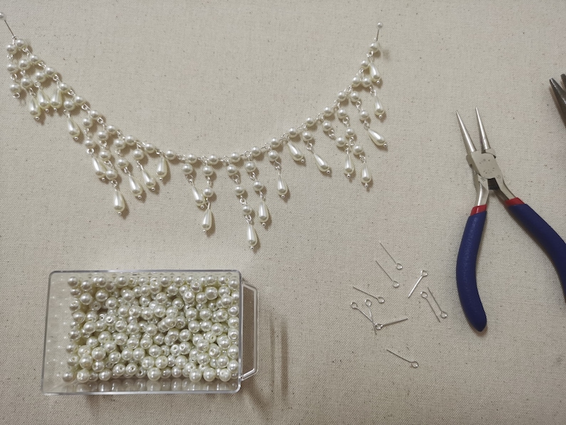 Op de afbeelding: Een close-up van een parelketting met een traanvormig ontwerp, een container met losse parels, een tang en een stapel springringen. De ketting is gemaakt van witte parels en een zilveren ketting.