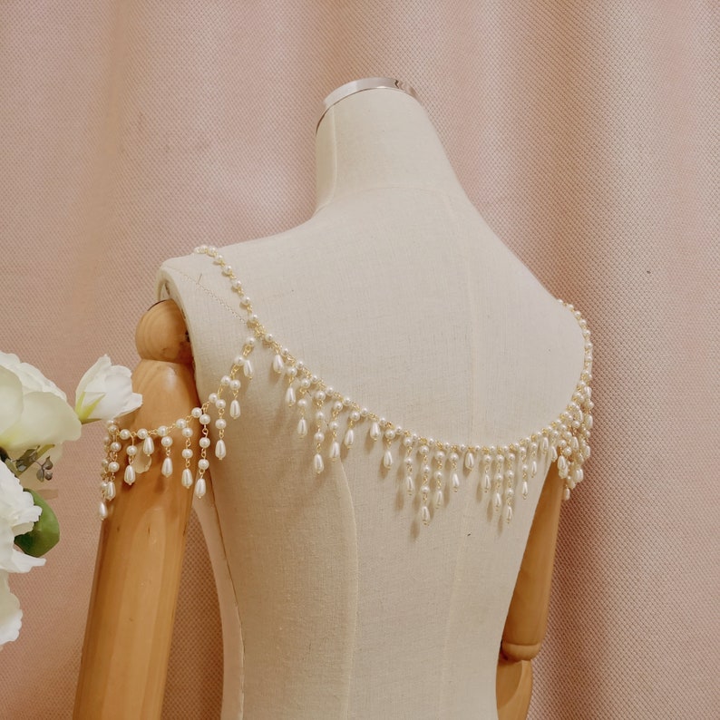 Op de afbeelding: Een schouderstuk van witte parels en een gouden ketting met hangende parels. Het stuk wordt getoond op een wit mannequins.