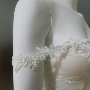 Könnte beinhalten: Nahaufnahme eines weißen Brautkleides mit zarten floralen Spitzendetails. Das schulterfreie Design verfügt über aufwendige Spitzärmel, die einen Hauch von Eleganz verleihen. Das Kleid wird auf einer Schaufenquin präsentiert.