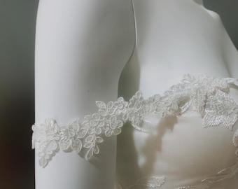 May include: Close-up of a white bridal gown with delicate floral lace detailing. The off-the-shoulder design features intricate lace sleeves, adding a touch of elegance. The dress is displayed on a mannequin.