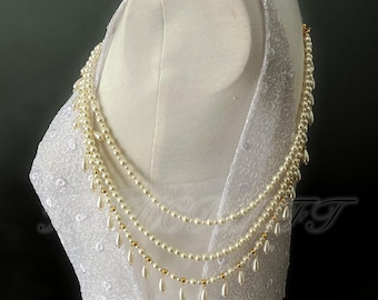 May include: An ivory pearl necklace with gold details and teardrop pendants. The necklace is draped over white, textured fabric, possibly a bridal gown. The design features multiple strands of pearls.