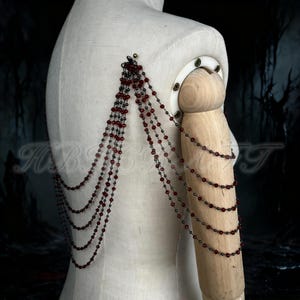 Puede incluir: Una pieza de joyería para hombro y brazo con hileras de cuentas rojas oscuras. Las cuentas están ensartadas en una cadena de metal oscuro, creando un efecto de capas. La joya está sujeta a un maniquí, mostrando su diseño y drapeado.