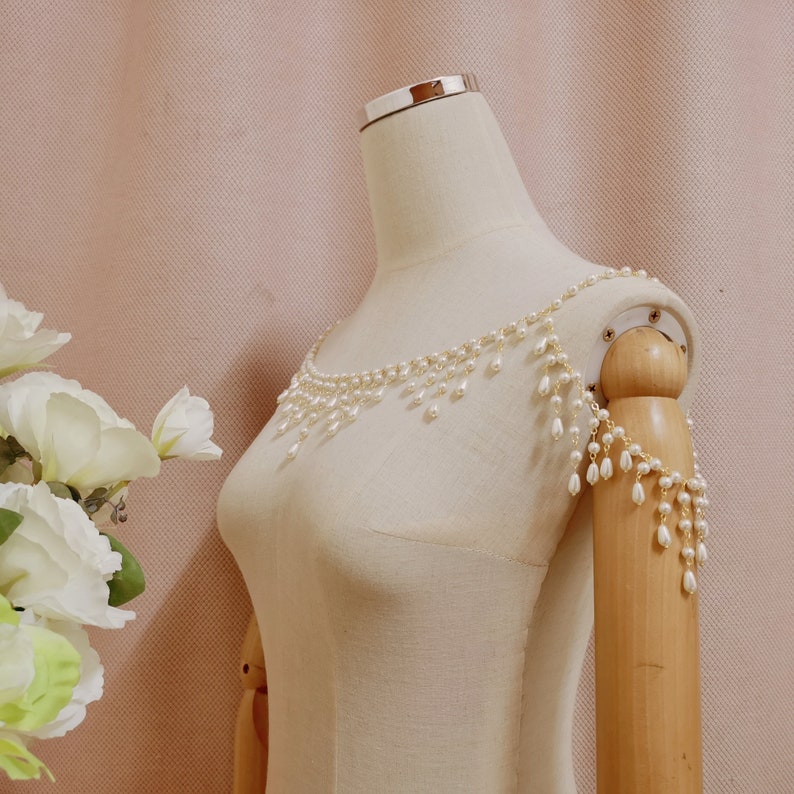 Op de afbeelding: Een witte parelketting met een delicate ketting en hangende parels. De ketting wordt getoond op een wit mannequin.