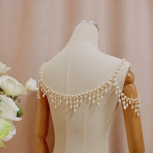 Op de afbeelding: Een schouderstuk van witte parels en een gouden ketting met hangende parels. Het stuk wordt getoond op een wit mannequin.