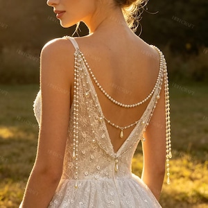 Può includere: Un abito in pizzo bianco con una profonda scollatura sulla schiena, decorato con molteplici fili di perle bianche drappeggiate. I fili di perle scendono dalle spalle e dalla schiena, creando un effetto a strati con piccole perle a goccia.