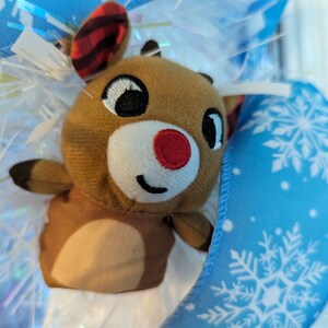 May include: A brown plush reindeer with a red nose and a red and black plaid hat. The reindeer is nestled in a blue and white snowflake patterned fabric.