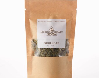 Puede incluir: Una bolsa de papel marrón con una ventana transparente que muestra hierba de Skullcap verde seca. La bolsa tiene una etiqueta blanca con el texto "BOTANICALS - HERBS - POWDERS - RESINS" y un logotipo que dice "MOON MYSTICRAFT". La etiqueta también dice "SKULLCAP 8g | 0.28 oz."