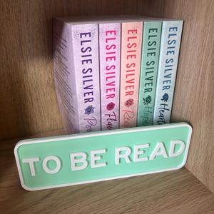 Op de afbeelding: Een verzameling boeken van Elsie Silver, met titels als "Heart", "Hope" en "Reck", uitgestald op een plank. Een mintgroen bord met de tekst "TO BE READ" staat voor de boeken, wat een leeshoekthema creëert.