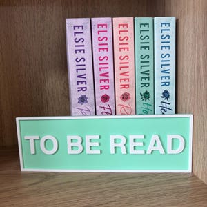 Op de afbeelding: Vijf boeken van Elsie Silver met bloemmotieven, achter een mintgroen bord met de tekst "TO BE READ". De boeken staan op een houten plank.