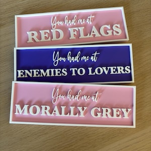 Pode incluir: Três placas retangulares com bordas e texto brancos. A placa superior é rosa com as palavras "You had me at RED FLAGS" em branco. A placa do meio é azul com "You had me at ENEMIES TO LOVERS". A placa inferior é rosa com "You had me at MORALLY GREY".