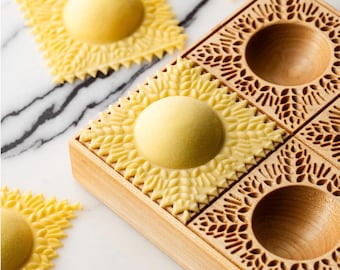 May include: A wooden ravioli mold with four sections, each with a decorative design. The mold is used to create decorative ravioli with a unique shape. There are four pre-made ravioli on a marble surface.