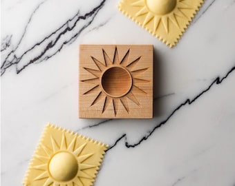 May include: A wooden ravioli stamp and two sun-shaped ravioli on a marble surface. The stamp is square with a sunburst design. The ravioli are yellow with a sun design and a scalloped edge.
