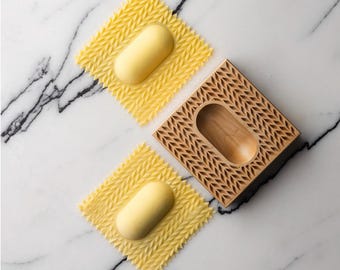 May include: A wooden ravioli mold with a textured design. Two yellow ravioli are shown, one in the mold and one on a white marble surface.