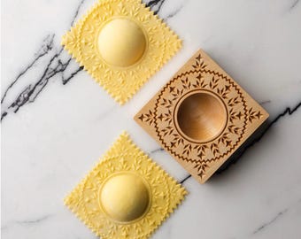 May include: Two yellow pasta squares with intricate designs and a wooden mold for making ravioli. The mold has a circular cutout in the center.