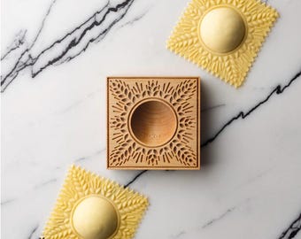 May include: A wooden ravioli mold with a circular indentation in the center. Two yellow ravioli are shown, one on each side of the mold. The ravioli have a decorative design of wheat stalks.
