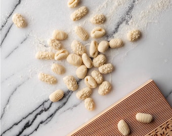 May include: A close-up of a wooden gnocchi board with a patterned surface. The board is surrounded by small, white gnocchi on a marble surface.