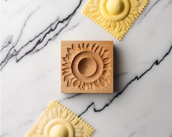 May include: A wooden mold for making pasta in the shape of a sunflower. Two pieces of pasta are shown, one on each side of the mold, with the same sunflower design.