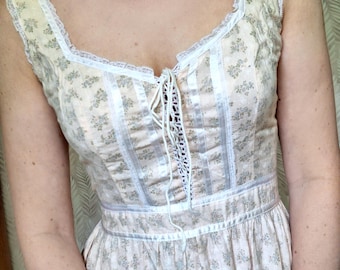 Puede incluir: Un vestido blanco sin mangas con estampado floral y ribete de encaje. El vestido tiene un corpiño con cordones y una falda fruncida.