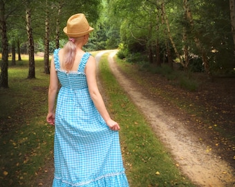 Puede incluir: Una mujer con un sombrero de paja camina por un sendero en un bosque. Lleva un vestido de cuadros azules y blancos con un ribete de encaje blanco.