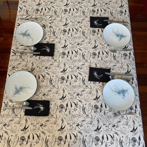 May include: A white tablecloth with a black and white bird pattern. Four place settings are set with white plates, silver cutlery, and black napkins.