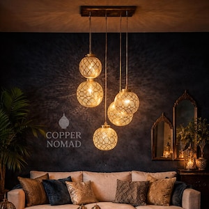 Peut inclure: Un salon avec un canapé beige, des coussins décoratifs et une table basse en bois sculpté. Des lampes suspendues dorées, sphériques et ornées de motifs pendent du plafond. Le texte "COPPER NOMAD" est visible.