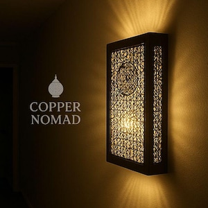 Peut inclure: Applique murale rectangulaire avec un cadre en bois foncé et un motif en métal ajouré. La lumière chaude émise par l'applique crée des motifs sur le mur. Le texte "COPPER NOMAD" est visible.