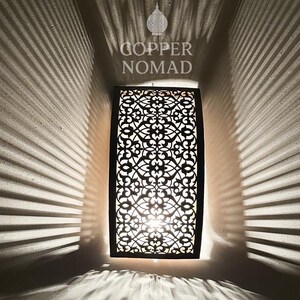 Peut inclure: Applique murale rectangulaire noire avec un motif ajouré complexe, projetant des ombres. La lumière vive émane de l'intérieur. Le texte "COPPER NOMAD" est au-dessus de l'applique.