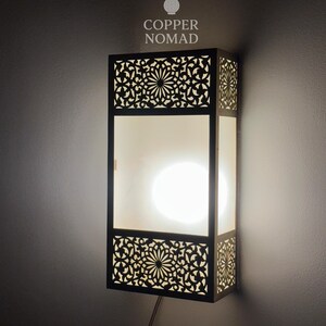 Peut inclure: Applique murale rectangulaire avec un design ajouré décoratif. L'applique a un cadre sombre avec des motifs floraux complexes et une source de lumière vive et diffuse. Le texte "COPPER NOMAD" est en haut.