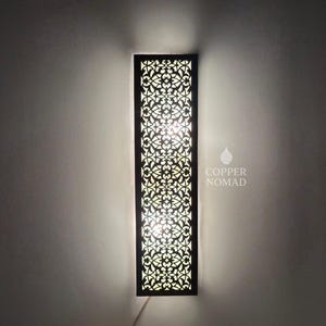 Pode incluir: Arandela de parede retangular com moldura escura e um intrincado design vazado floral. A arandela é iluminada por dentro, projetando um brilho quente. O texto "COPPER NOMAD" é visível.