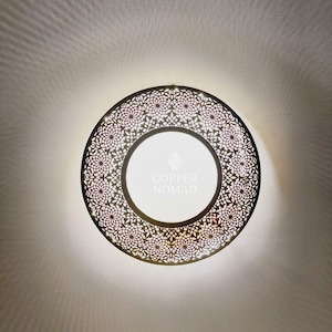 Puede incluir: Una lámpara redonda y decorativa con un borde detallado con motivos florales. El centro de la lámpara es blanco, con el texto "COPPER NOMAD". La lámpara está montada en una pared blanca.