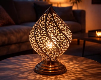 May include: Ornate, teardrop-shaped table lamp with a bronze finish. The lamp features intricate cut-out patterns that cast decorative shadows. The lamp sits on a round base and is illuminated, creating a warm ambiance. The lamp is on a wooden table.
