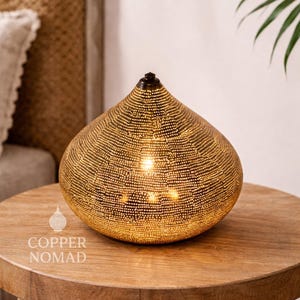 Op de afbeelding: Een decoratieve, druppelvormige lamp met een getextureerd, geperforeerd oppervlak. De lamp is gemaakt van goudkleurig metaal en staat op een houten tafel. De lamp is van binnenuit verlicht en geeft een warme gloed. De tekst "COPPER NOMAD" is zichtbaar.