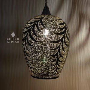 May include: A black pendant lamp with a leaf-like pattern of small, evenly spaced holes. The lamp is suspended from a dark cord and has a teardrop shape. The light shines through the holes, creating a warm glow. The text "COPPER NOMAD" is visible.