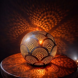 Puede incluir: Lámpara decorativa esférica marrón oscuro con un diseño de pequeños agujeros espaciados. La lámpara emite un brillo naranja cálido, proyectando intrincados patrones de luz sobre la superficie circundante. La lámpara mide aproximadamente 20 cm de diámetro.