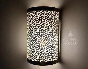May include: A wall-mounted, cylindrical lamp with a black metal frame and a patterned, perforated shade. The shade's design allows light to filter through, creating a warm glow. The text "COPPER NOMAD" is visible on the right side.