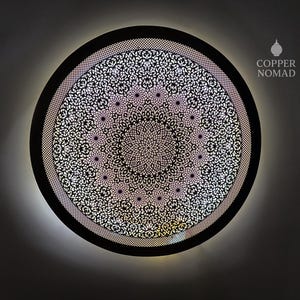 Puede incluir: Una pieza de arte mural redonda de metal negro con un detallado patrón floral blanco. El diseño está iluminado desde atrás, creando un brillo suave. El texto "COPPER NOMAD" está en la esquina superior derecha.