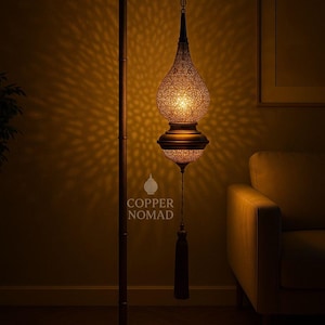 Op de afbeelding: Een bronzen vloerlamp met een sierlijke, patroonvormige kap. De lamp heeft een lange steel en een decoratieve kwast. Het ontwerp van de lamp werpt een warm, patroonvormig licht op de muur. De basis van de lamp is vierkant.