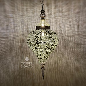 Peut inclure: Une lampe suspendue en métal orné, en forme de goutte d'eau. La lampe présente des motifs ajourés complexes qui projettent des ombres décoratives. La lampe a une finition sombre. Le texte "COPPER NOMAD" est visible.