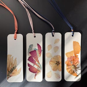 May include: Four handmade bookmarks with pressed flowers and colorful ribbons. Each bookmark features a unique floral arrangement in shades of orange, pink, and beige, set against a white background. The ribbons are in shades of peach, pink, and navy.