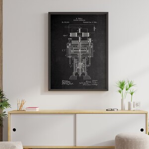 May include: Black and white illustration of a patent for an electric generator, designed by Nikola Tesla. The patent number is 514,168 and the date is January 5, 1894. The illustration is framed and hanging on a white wall above a white cabinet with a wooden top.