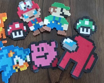 May include: A collection of pixel art figures featuring characters from popular video games. Includes Mario, Luigi, Crash Bandicoot, Sonic the Hedgehog, a red Among Us character, and various mushroom designs. The figures are made of colorful plastic beads.