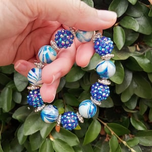 May include: A blue and white beaded bracelet with a mix of textures. The bracelet features large, marbled beads and smaller, sparkling, crystal-covered beads. Silver-toned spacers separate the beads. The bracelet is held against a backdrop of green foliage.