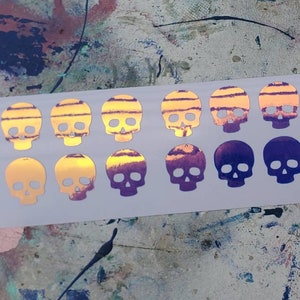 Pode incluir: Uma folha de 18 adesivos de caveira hologr&aacute;ficos iridescentes. As caveiras est&atilde;o em um degrad&ecirc; de roxo e amarelo.
