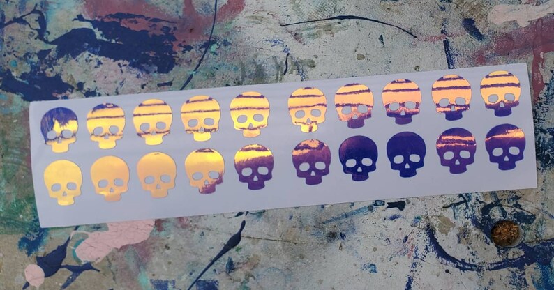 Pode incluir: Uma folha de 18 adesivos de caveira hologr&aacute;ficos iridescentes. As caveiras est&atilde;o em um degrad&ecirc; de roxo e amarelo.