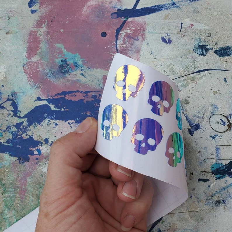Pode incluir: Uma folha de adesivos de caveiras iridescentes. As caveiras s&atilde;o em tons de azul, roxo e verde. Os adesivos est&atilde;o sobre um fundo branco.