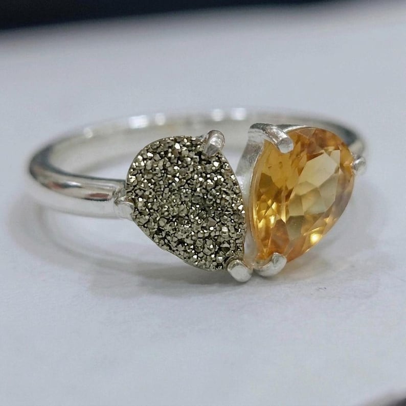 Op de afbeelding: Een zilveren ring met twee stenen naast elkaar: de ene is een getextureerde, metallic grijze druzy-steen met een sprankelend oppervlak, en de andere is een gladde, gefacetteerde, peervormige gele citrien, beide gezet in een zilveren band.
